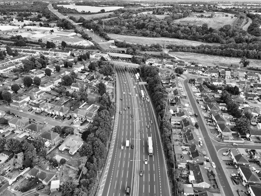 Aerial view in black and white showing a multi-lane highway with various vehicles, including cars, vans, and trucks, moving along the road. The highway passes through a suburban area with rows of sing